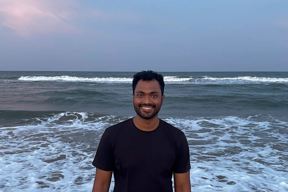 Shreyas standing in the surf at dusk, waves breaking behind him under a pink sky.